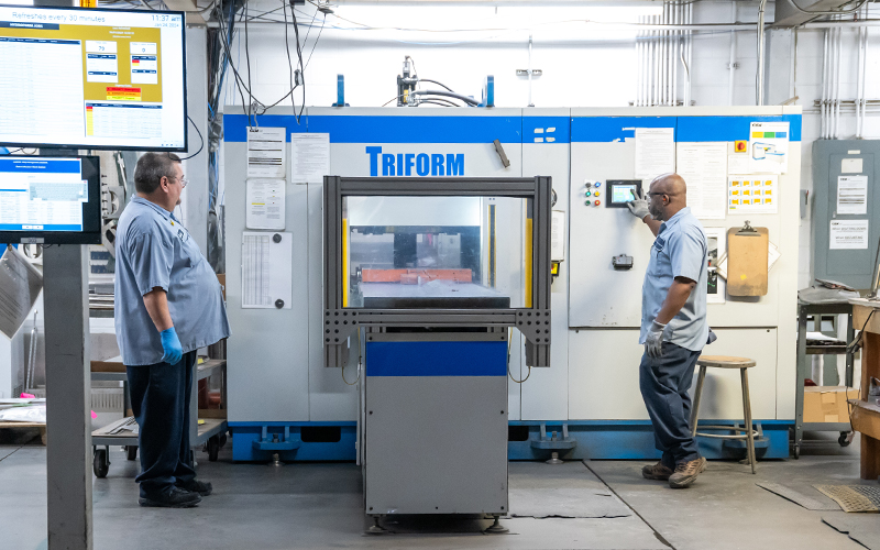 Two Re:Build CDI employees standing in front of a Triform Hydroforming machine working on a piece of equipment